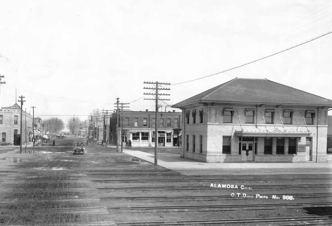 Black and white photograph of a railroad crossing with multiple rails, in a town, next to roads