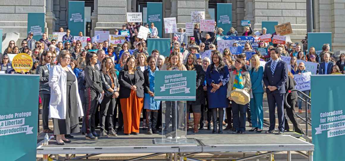 Photo of a large group of people holding up signs on the Colorado state capitol steps behind a podium and speaker