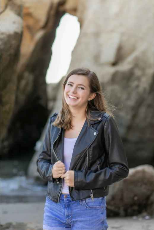 Headshot photo of Maya Walthall, smiling, standing on a beach in front of a rock formation
